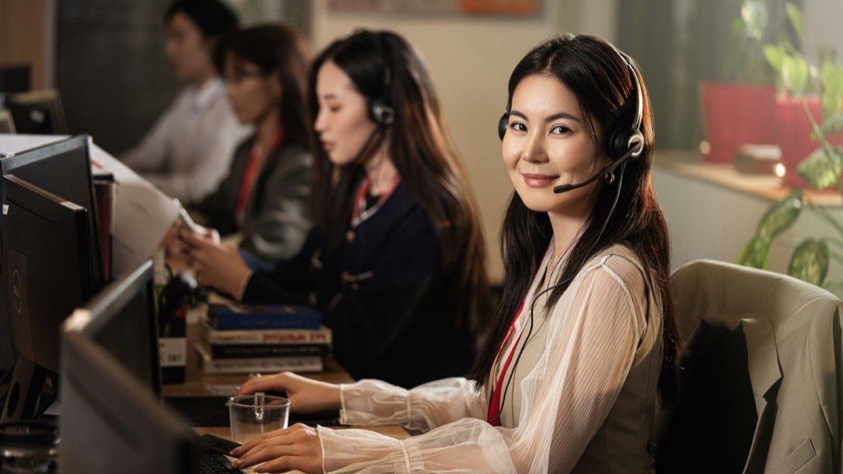 Conseillère souriante avec un casque téléphonique à son poste de travail, collègues en ligne derrière elle