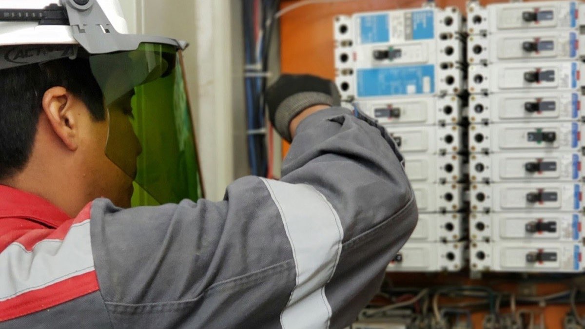 Électricien de dos, casque et visière de protection, intervenant sur une armoire de disjoncteurs avec un multimètre
