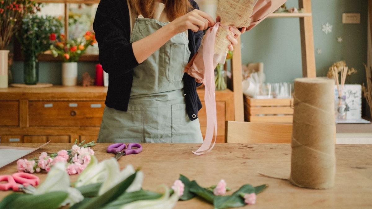 Fleuriste en tablier qui noue un ruban rose autour d’un bouquet sur un établi en bois