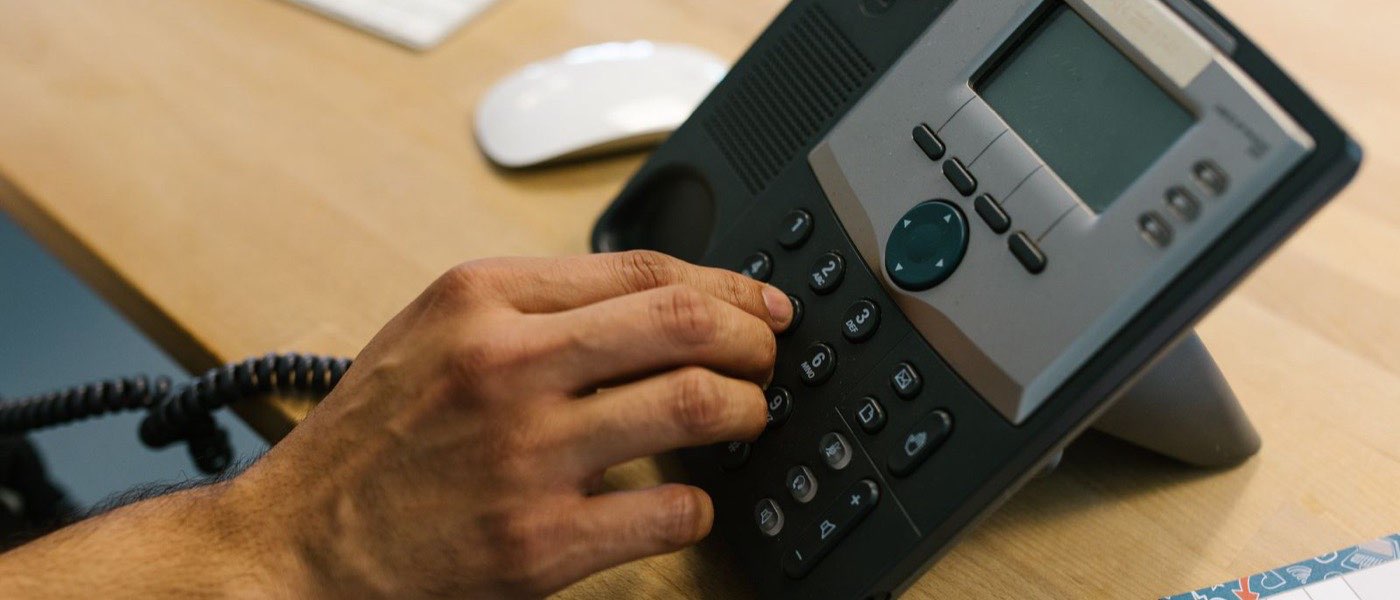 Main qui compose un numéro sur un téléphone fixe de bureau posé sur une table en bois